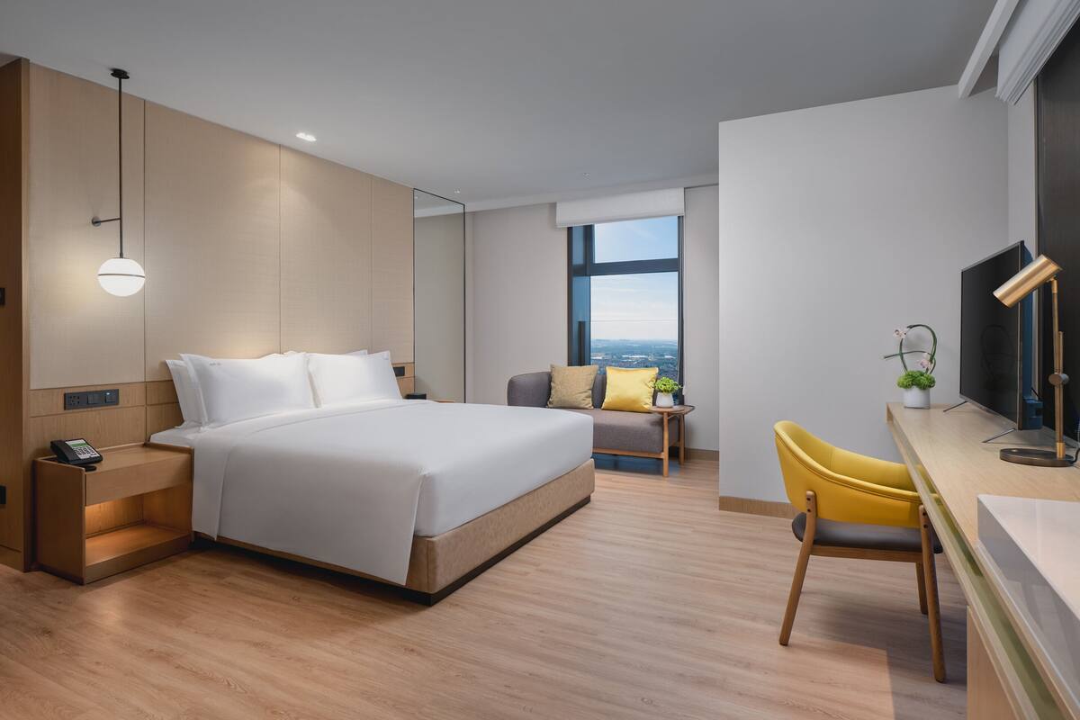 Una habitación de hotel moderna con cama, mesita de noche, lámpara, silla, escritorio, televisor y un ventanal con vistas a la ciudad.