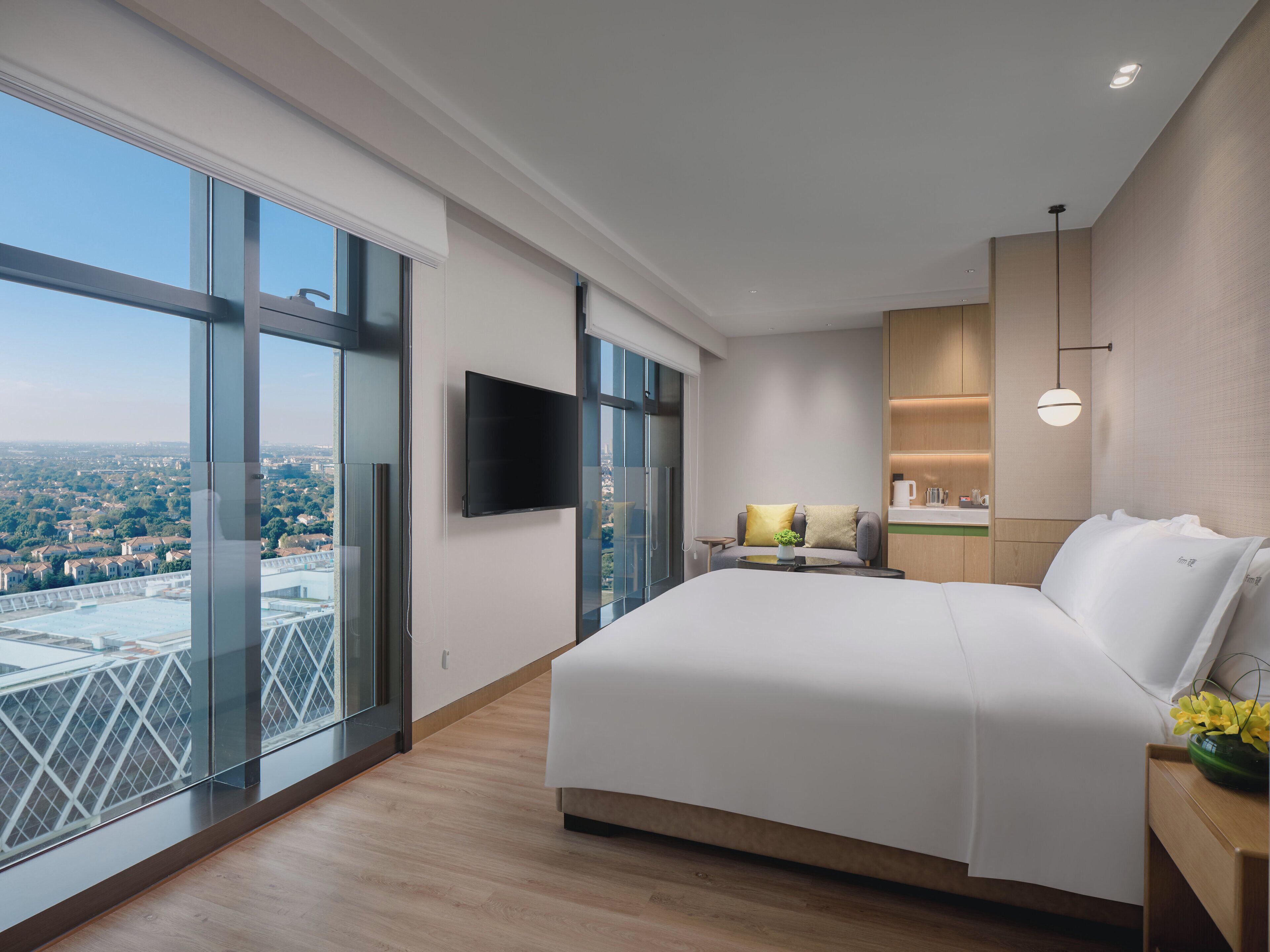A modern hotel room with a large bed, a flat-screen TV, a small table with a lamp, and a view of a cityscape through floor-to-ceiling windows.
