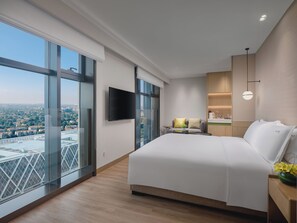 Ein modernes Hotelzimmer mit einem großen Bett, einem Flachbildfernseher, einem kleinen Tisch mit einer Lampe und Blick auf eine Stadtlandschaft durch raumhohe Fenster.