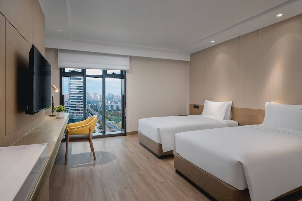 Una habitación de hotel moderna con dos camas, un escritorio, una silla y un ventanal con vistas a la ciudad.