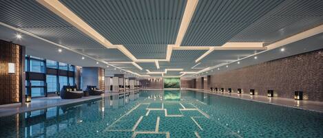 A large indoor swimming pool with a geometric tile pattern, modern ceiling design, and seating areas along the sides.