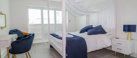 A bedroom with a canopy bed, a desk with a chair, a bedside table with a lamp, and a window with blinds.