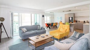 A modern living room with a sofa, armchair, and coffee table. There is a dining area with colorful chairs and a kitchen in the background. - 30 Cliff Edge (Newquay)