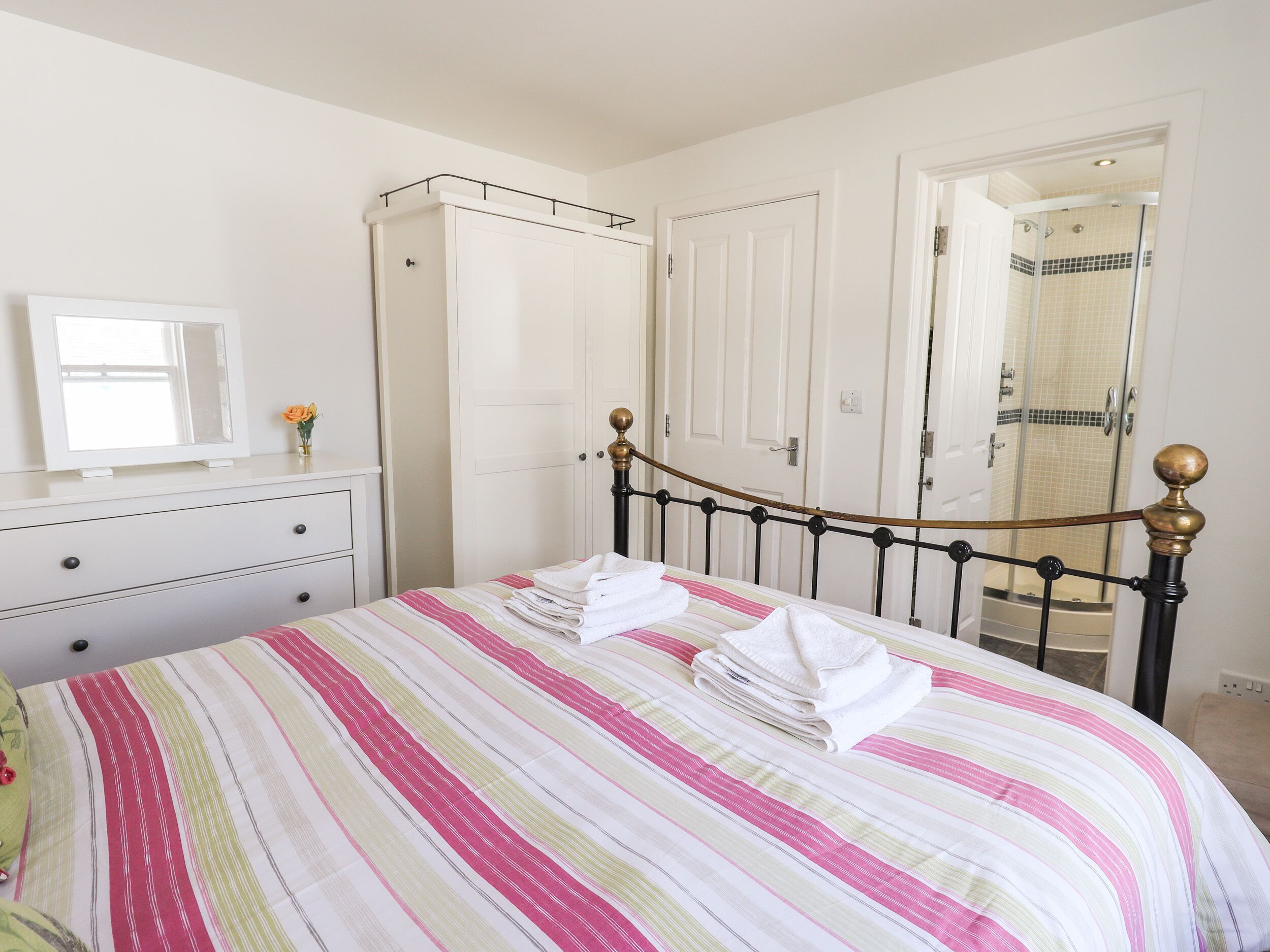A neatly made bed with striped bedding, a dresser with drawers, a mirror, and a door leading to a shower area.