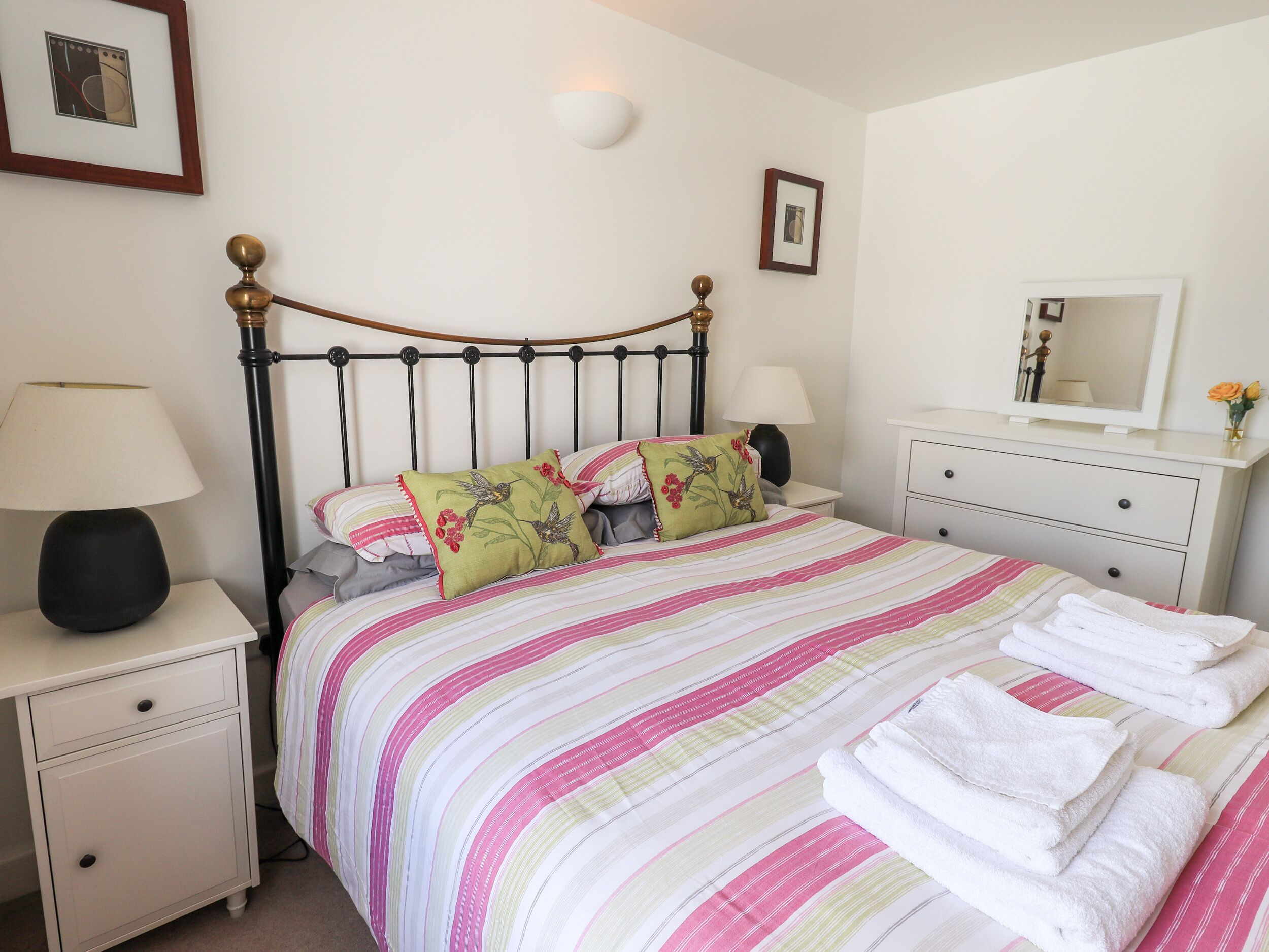 A neatly made bed with striped bedding, a bedside table with a lamp, a nightstand, a dresser with a mirror, and a framed picture on the wall.