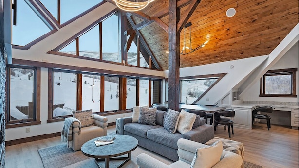A spacious living area with a high ceiling, large windows offering a view of a snowy landscape, and a cozy seating arrangement. - The Lodge at Mountain Village | Stunning Ski-In/Out Presidential Penthouse | A Nestique Property (Park City)