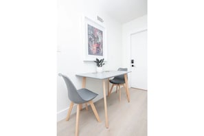 A minimalist dining area with a white table and two grey chairs, a framed abstract artwork on the wall, and a small potted plant on the table.