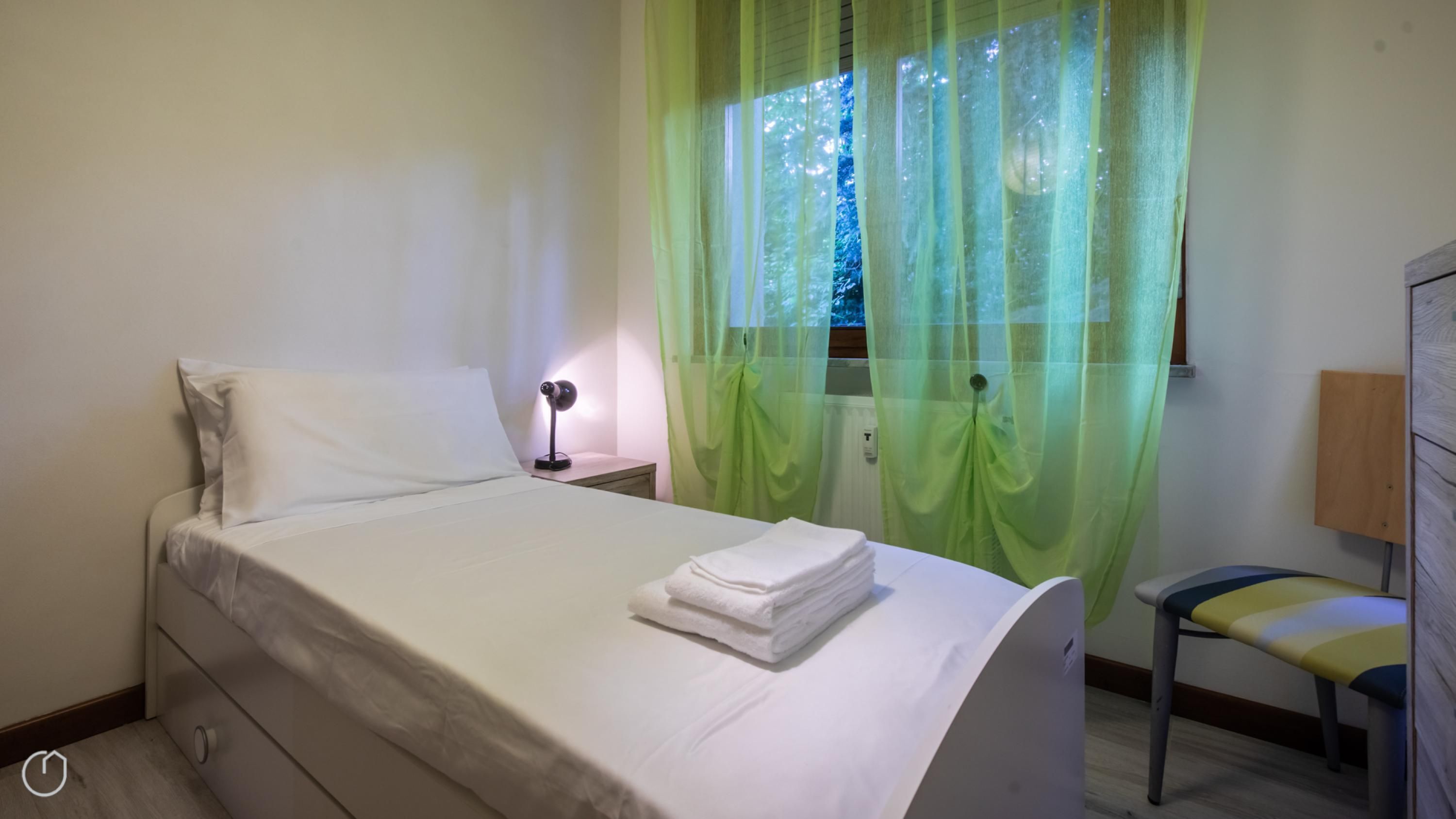 A neatly made bed with white linens, a bedside lamp, a chair, and sheer green curtains over a window.