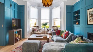 A living room with teal walls, a large window, a wooden ceiling light, and a television on a stand. - Exceptional Renovated Period Home in Clifton (Bristol)