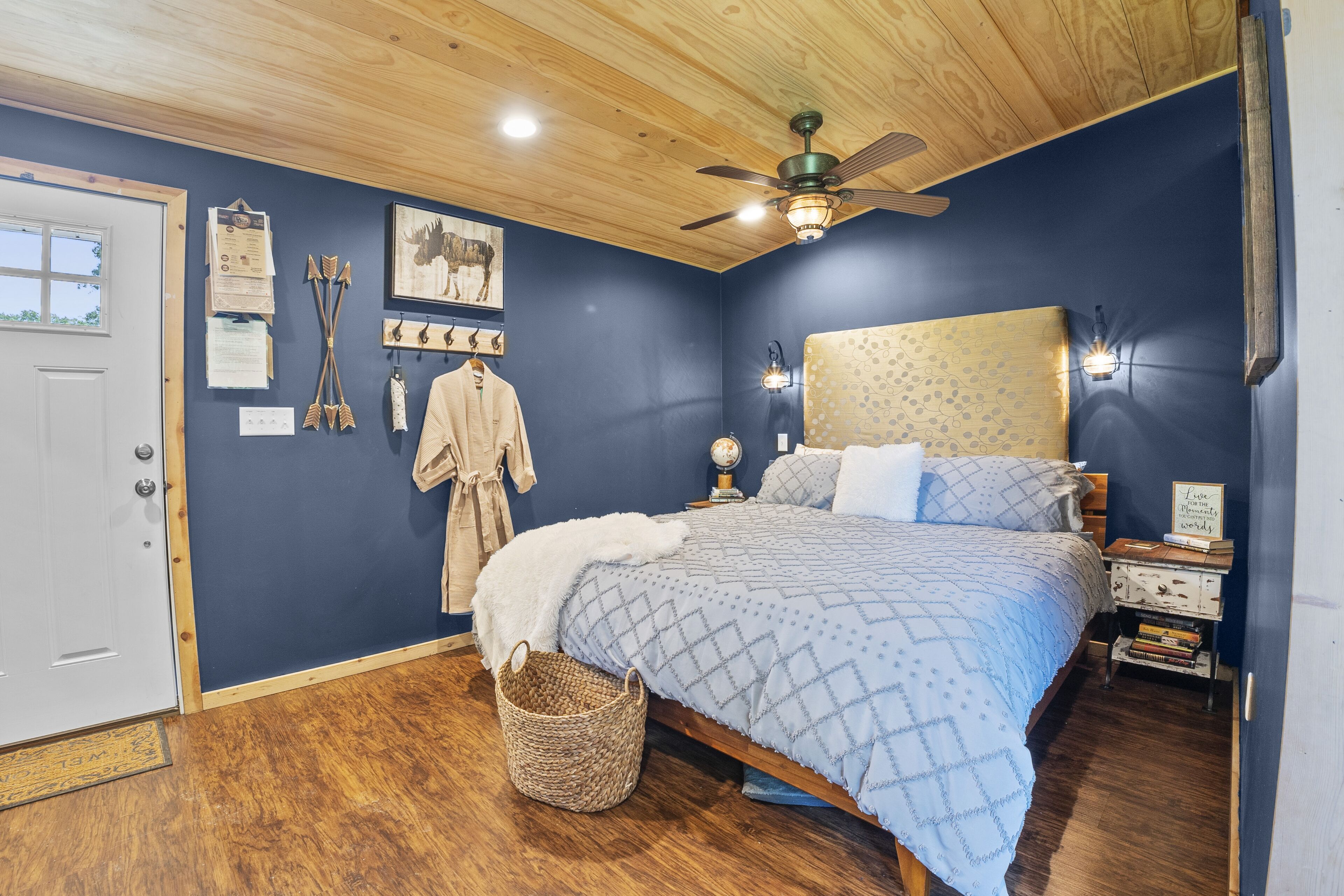 A bedroom with a bed, wooden ceiling, a ceiling fan, a nightstand, a lamp, and a wall with various items.