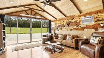 A spacious living room with a wooden ceiling, leather furniture, and a large window offering a view of the outdoors.