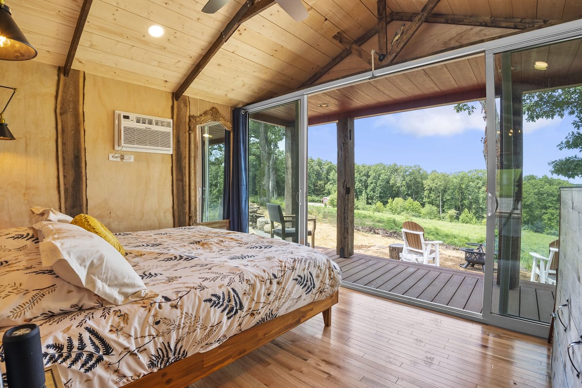 A bedroom with a large bed, wooden flooring, and a sliding glass door leading to an outdoor deck with chairs.