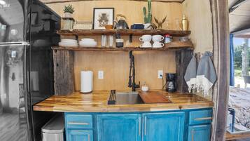 Cocina compacta con refrigerador, pileta y muebles azules. Se observan estantes con diversos objetos y una ventana con vista.