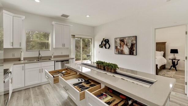 A modern kitchen with white cabinets, a large island with drawers, a window, and a painting on the wall. - Watching Wildlife: Private Oasis for 4 w/ Patio, BBQ & Parking. Step Inside! (Payson)