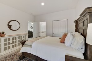 Une chambre à coucher avec un grand lit, une tête de lit en bois, une armoire blanche, un miroir et une porte donnant sur une autre pièce.