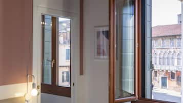 Un dormitorio con cama, lámpara de noche, ventana con vista a edificios y techo de madera.