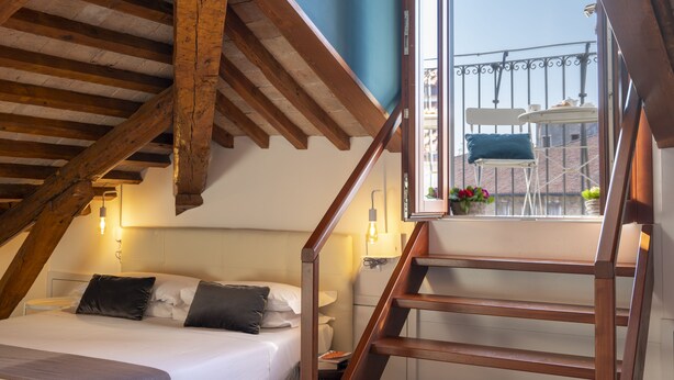 A bedroom with a sloped ceiling, wooden beams, a bed with pillows, a nightstand, a window with a view, and a staircase. - Fosca Venice Rooms (Venice)