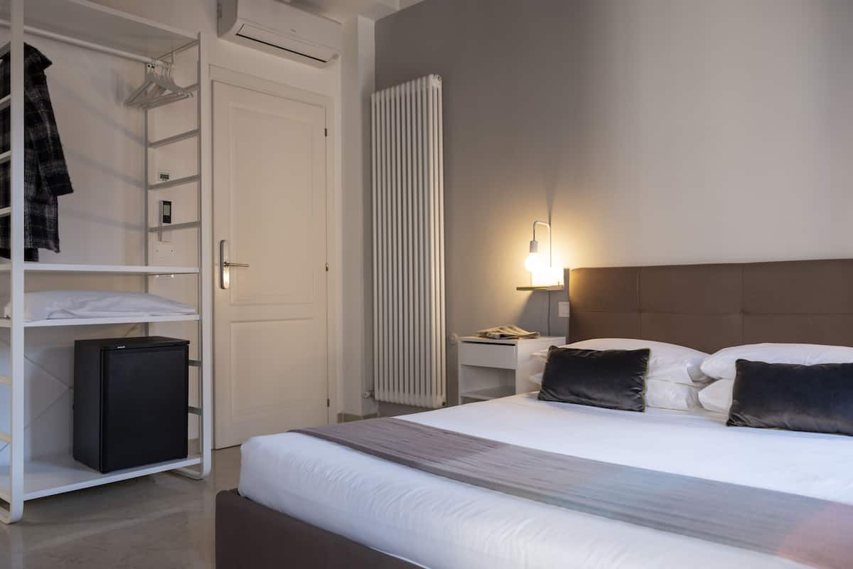 A modern hotel room with a large bed, a wardrobe, a bedside table with a lamp, and a radiator.