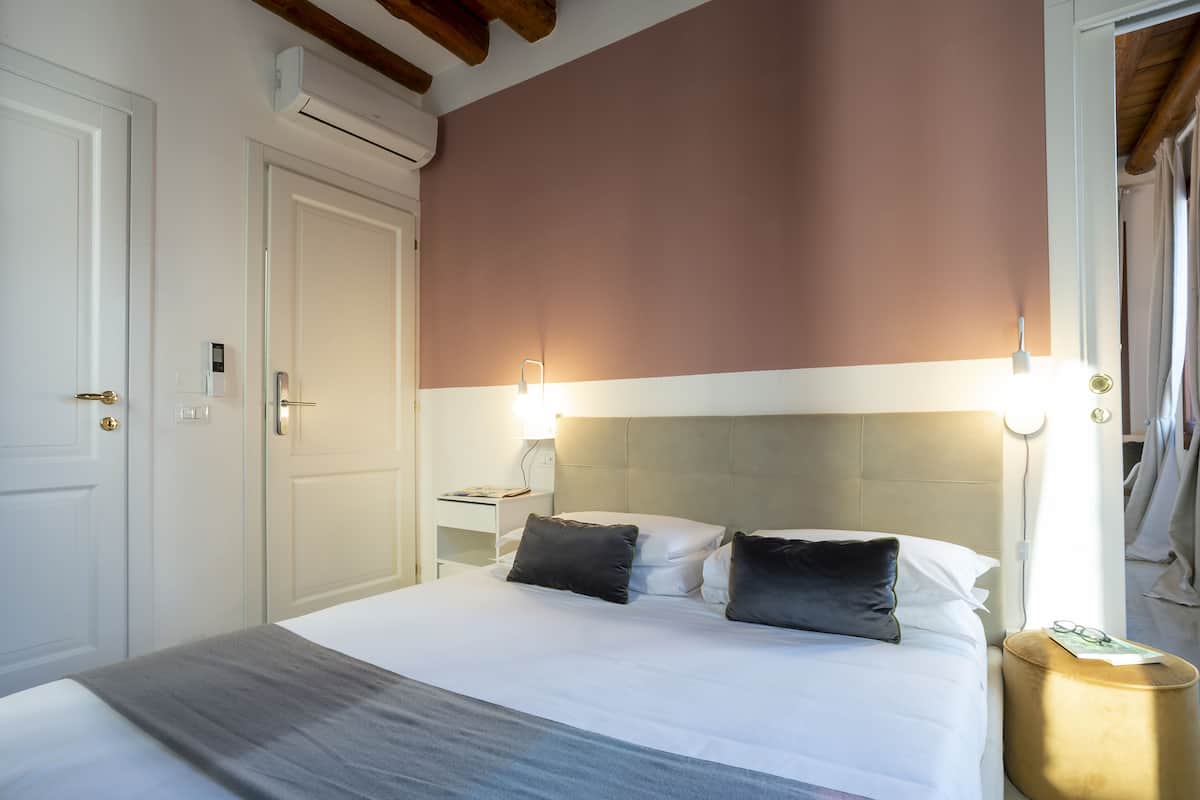 A neatly made bed with white linens and two dark pillows, a bedside table with a lamp, and a wall-mounted air conditioner.