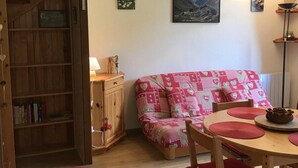 Un salon chaleureux avec une bibliothèque en bois, un canapé rouge, une table à manger avec des sets de table rouges et un tableau représentant un paysage de montagne accroché au mur.