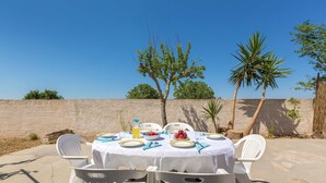 Zona pranzo all'aperto con tavolo apparecchiato per quattro, circondata da sedie in plastica bianca, e vista libera sul cielo e sugli alberi sullo sfondo.