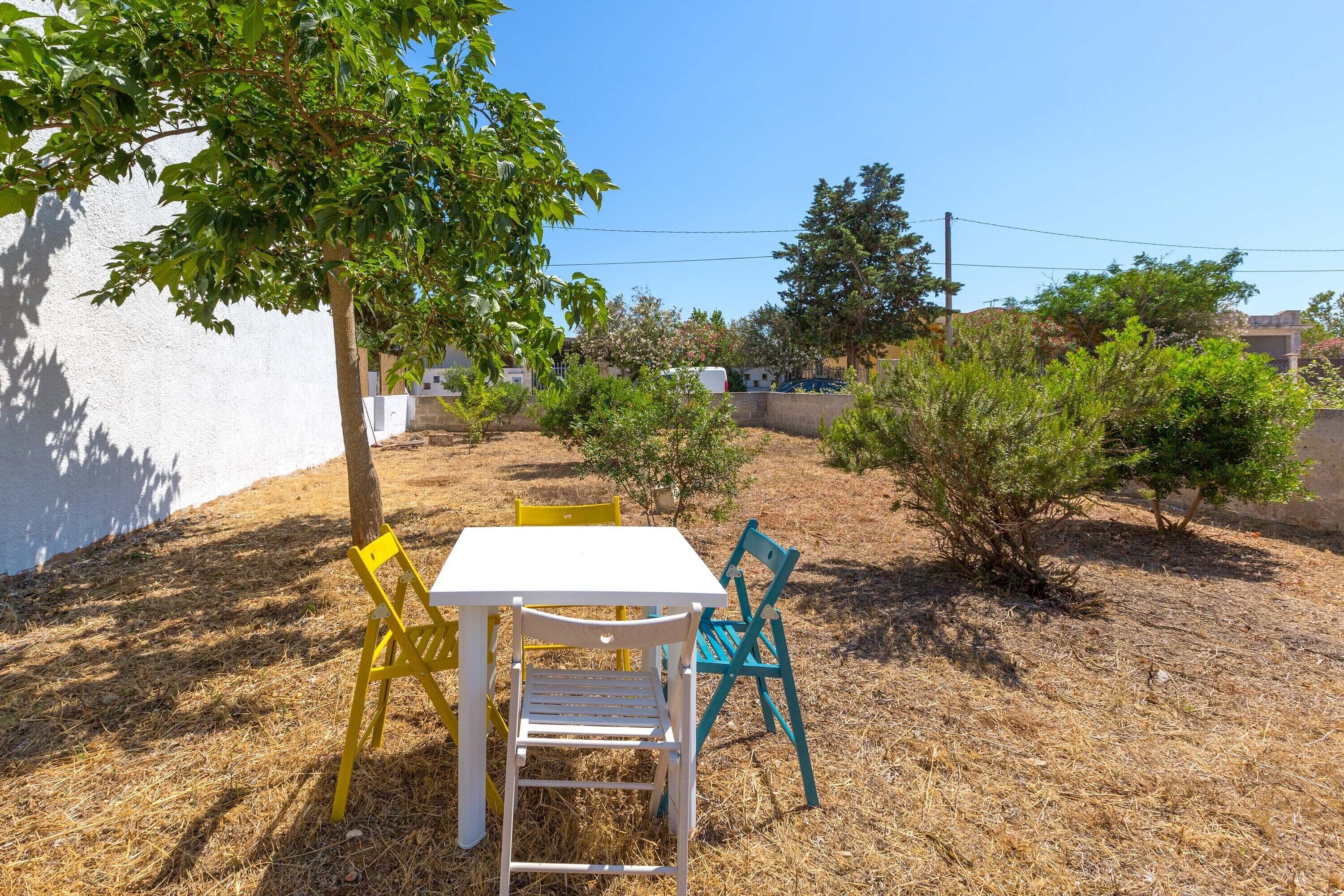 Un espace extérieur aménagé avec une table et des chaises blanches, entouré d’herbe sèche et de petits arbres.