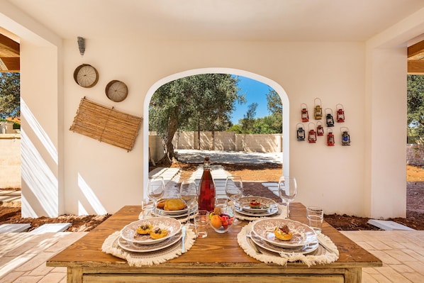 A dining table set with plates, glasses, and a bottle, with a view of trees and a stone wall through an archway.