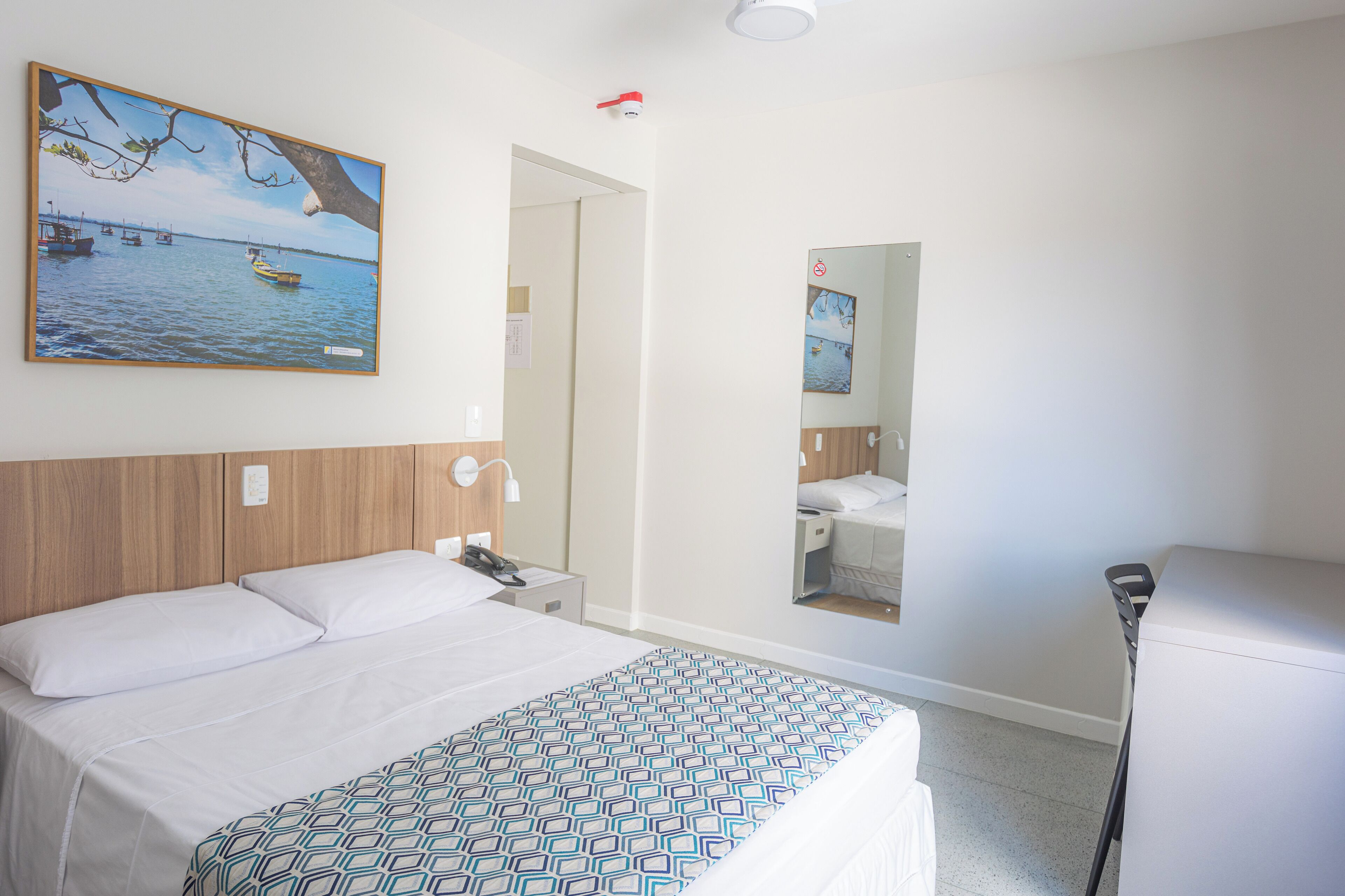 Habitación de hotel con cama, mesita de noche, espejo y un cuadro de una escena costera.
