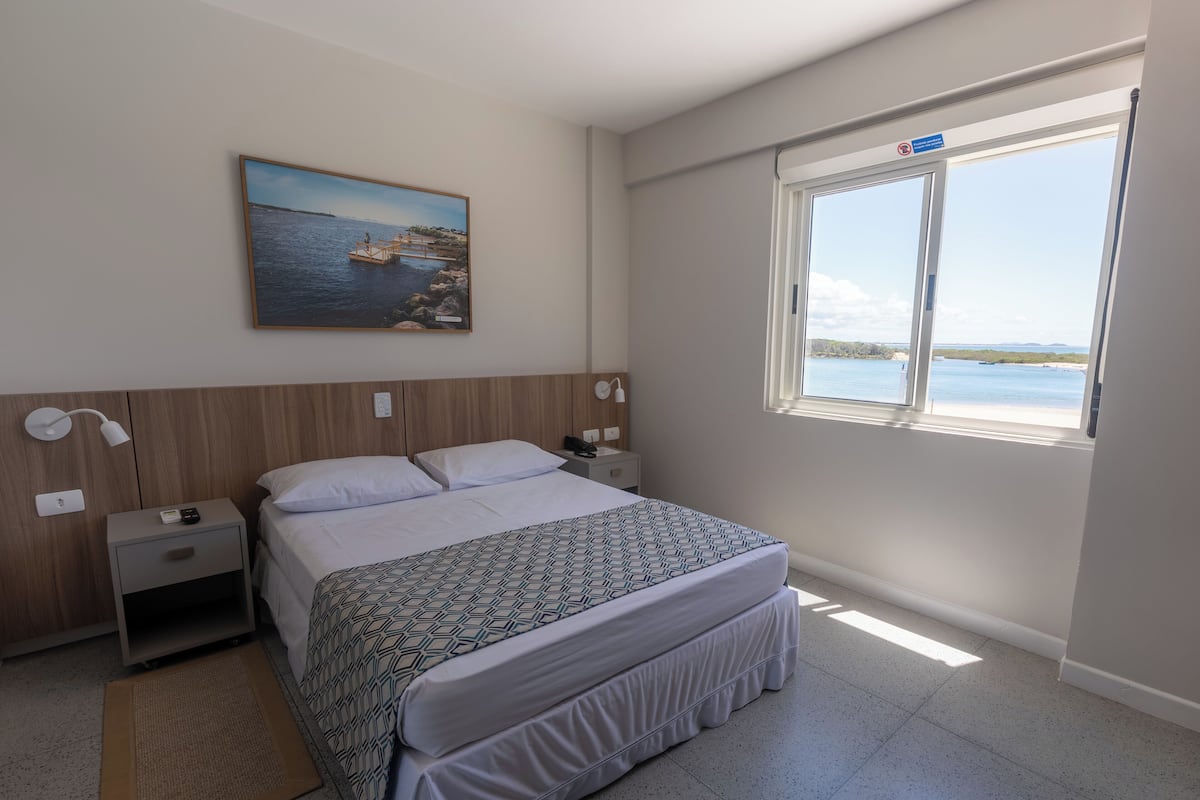 A bedroom with a bed, nightstands, a painting, and a window with a view of the sea.
