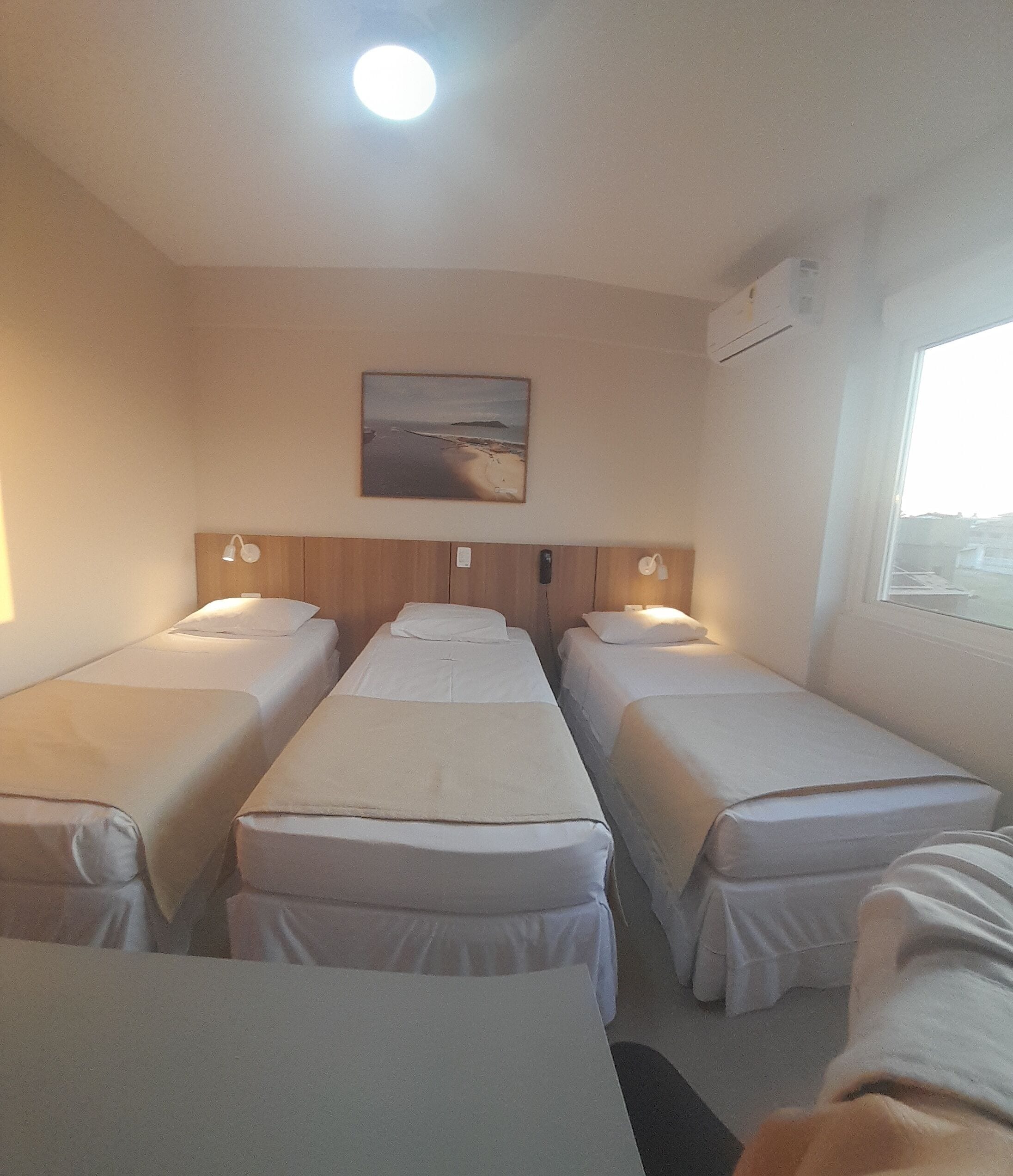 A room with three single beds, a wooden headboard, a painting of a beach, and a window with a view.