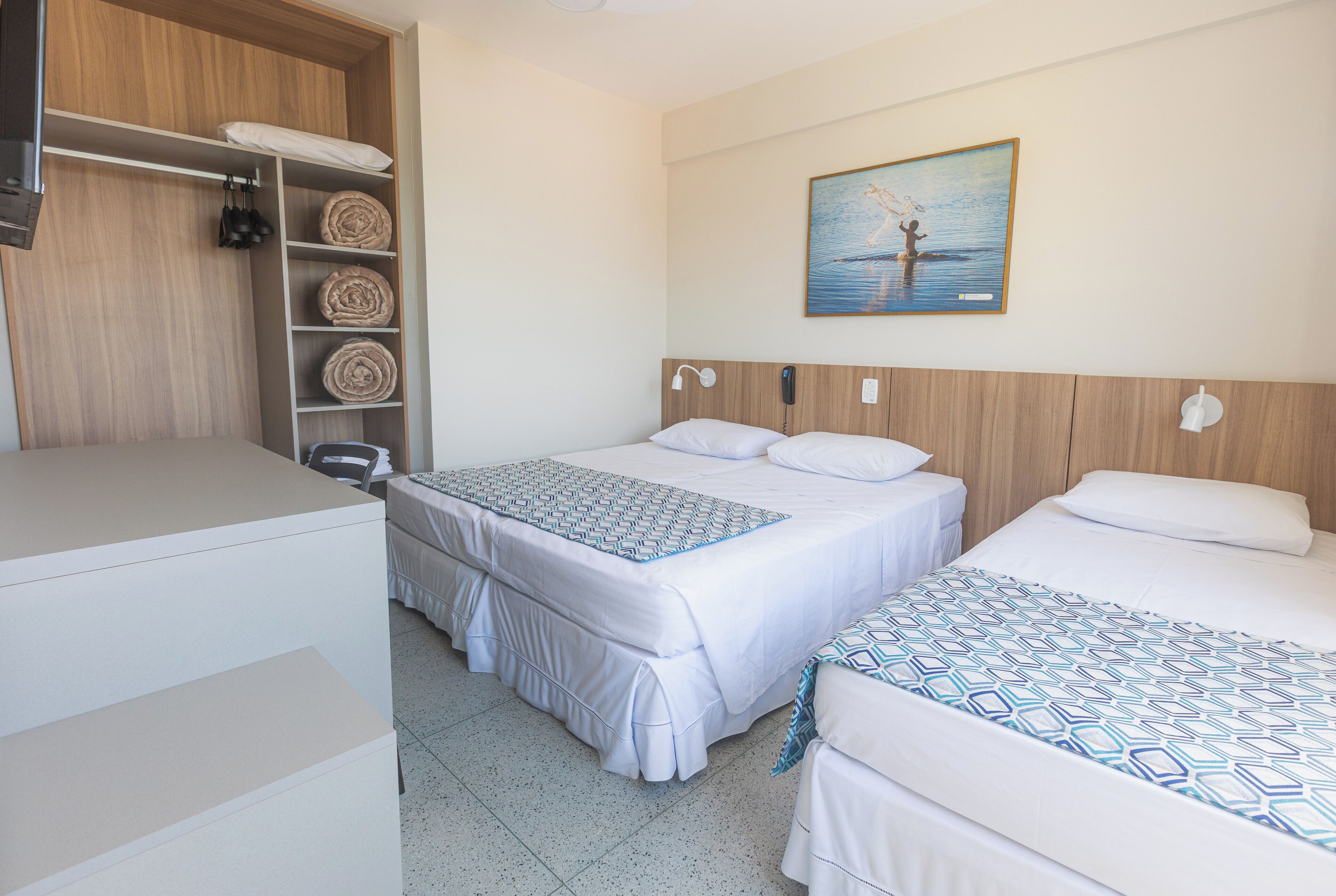 A hotel room with two beds, a wooden wardrobe, a desk, and a painting of a person by the sea.