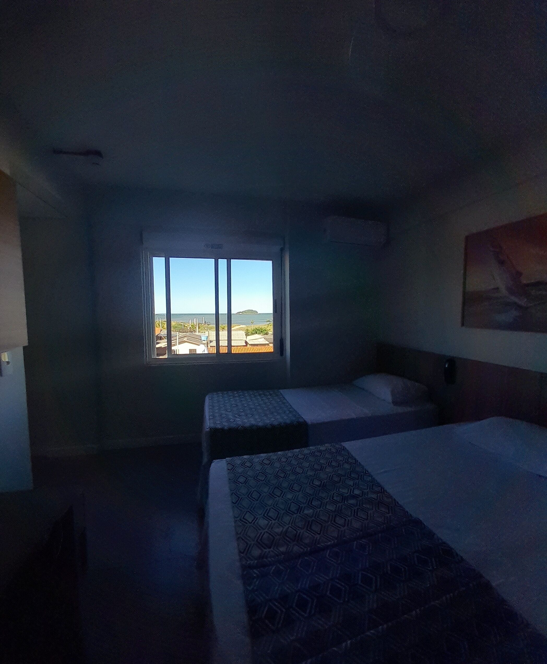 A dimly lit hotel room with two beds, a large window offering a view of buildings and a distant island, and a painting on the wall.