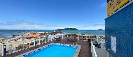 A pool area with a view of the sea, a deck with chairs, and a sign for 'Hotel Bandeirante da Barra'.