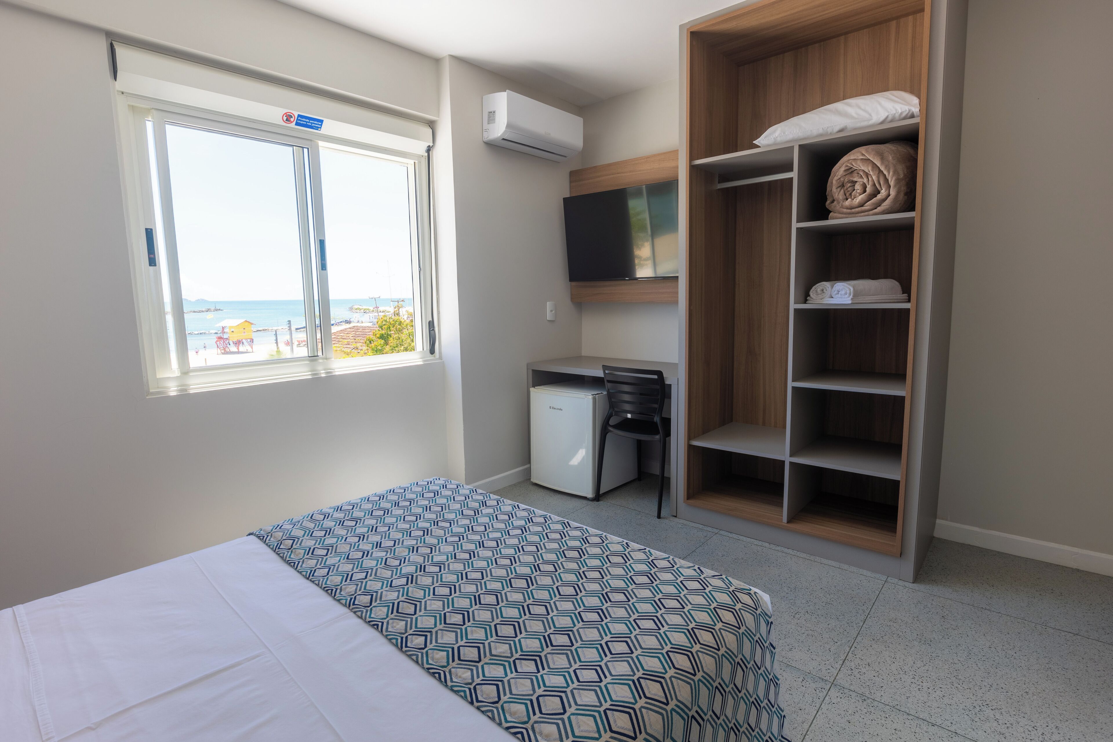 Quarto com cama, escrivaninha, cadeira, guarda-roupa, televisão e janela com vista para o mar.