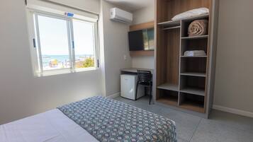 Quarto com cama, escrivaninha, cadeira, guarda-roupa, televisão e janela com vista para o mar.