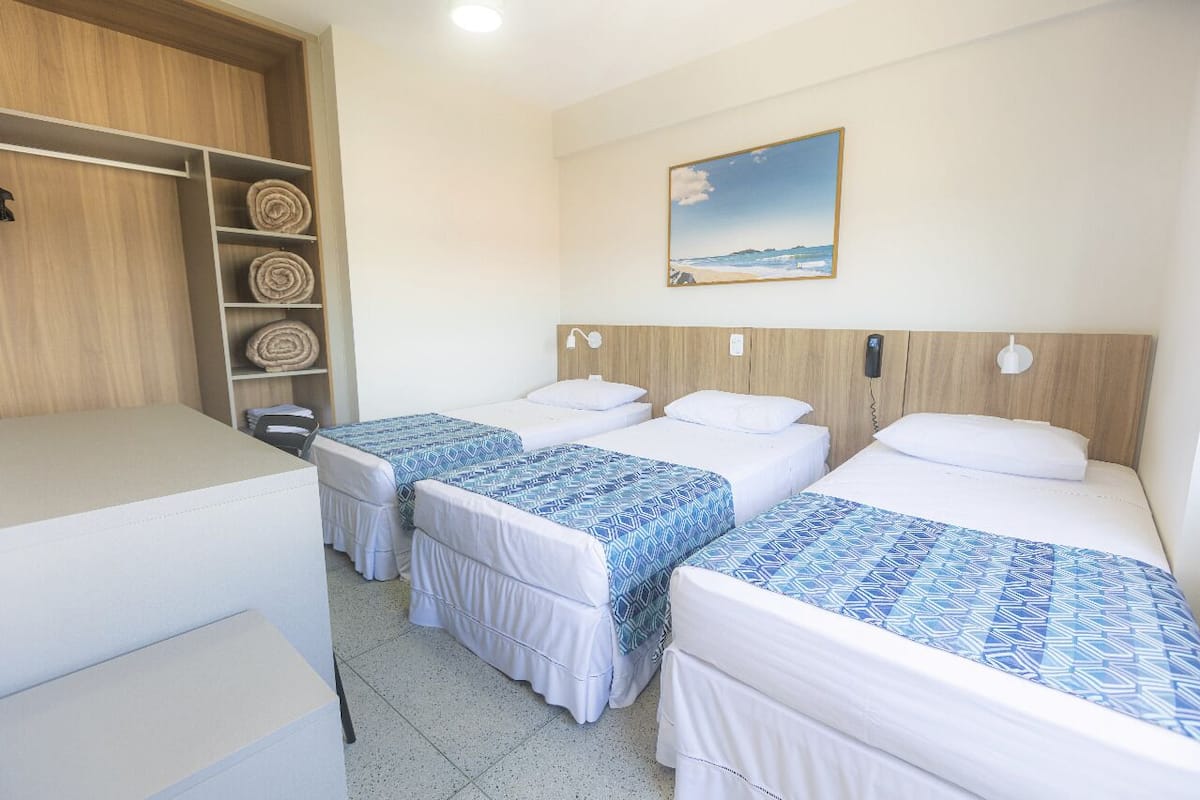 A hotel room with three beds, a wooden wardrobe, and a painting of a beach.