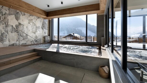 A modern spa area with a hot tub, wooden steps, and large windows offering a view of snowy mountains. - Wifi, fitness, swimming pool, sauna, hammam, balcony, tv, ski locker, 75m², Le Grand Bornand (Le Grand-Bornand)