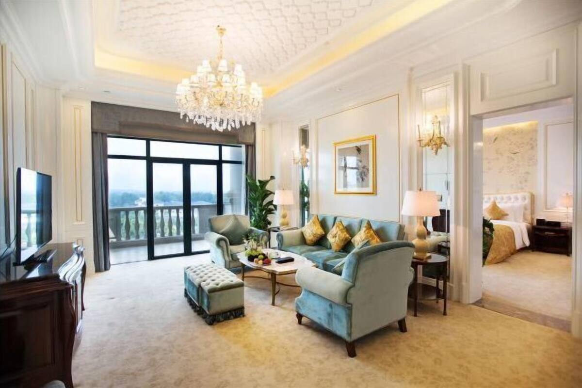 Ein großzügiger Wohnraum mit einer großen Couch, einem Couchtisch und einem Fernseher. Durch die großen Fenster ist ein Balkon sichtbar.