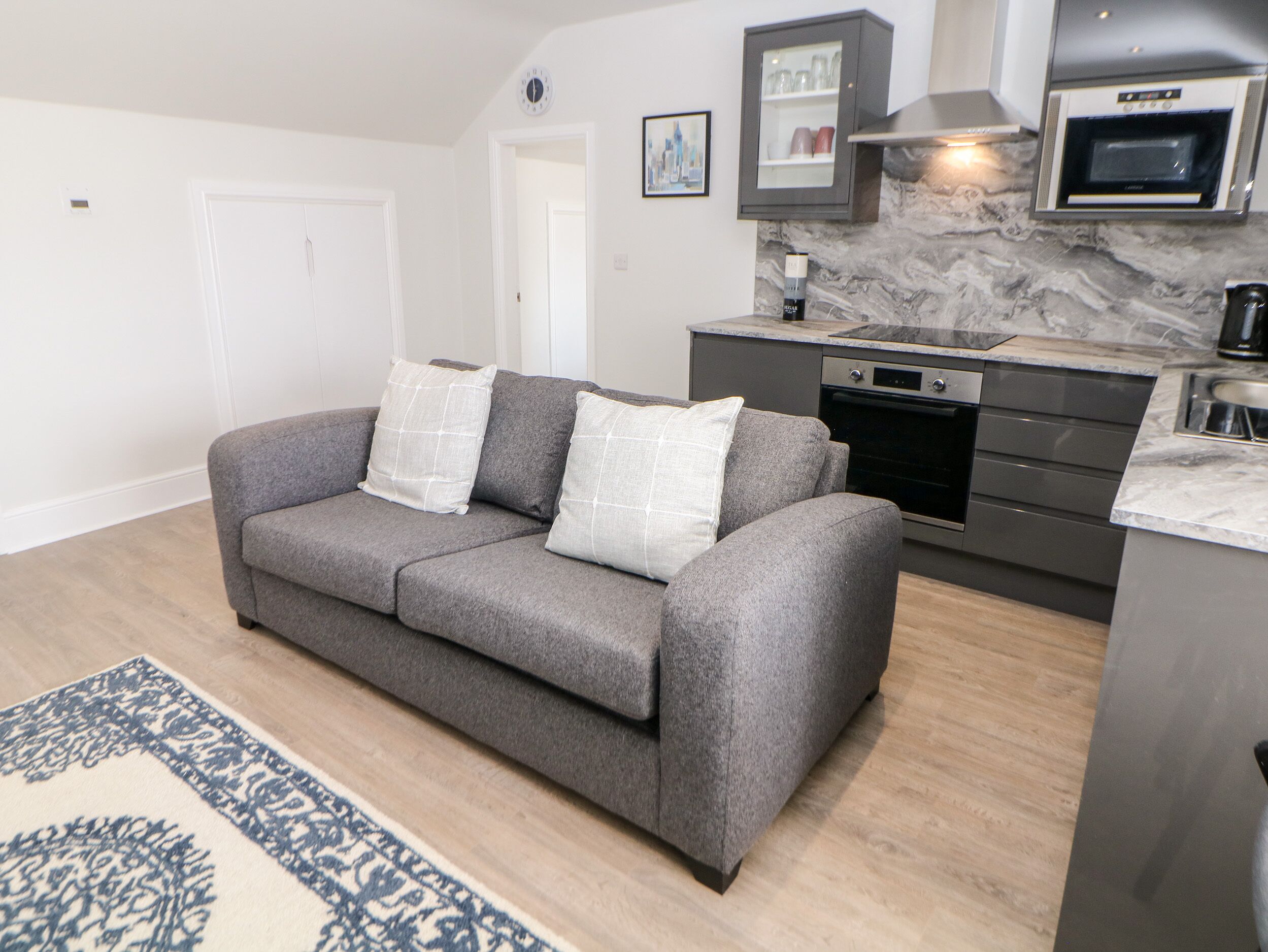 A modern kitchen with a built-in oven, a grey sofa with white cushions, and a patterned rug.