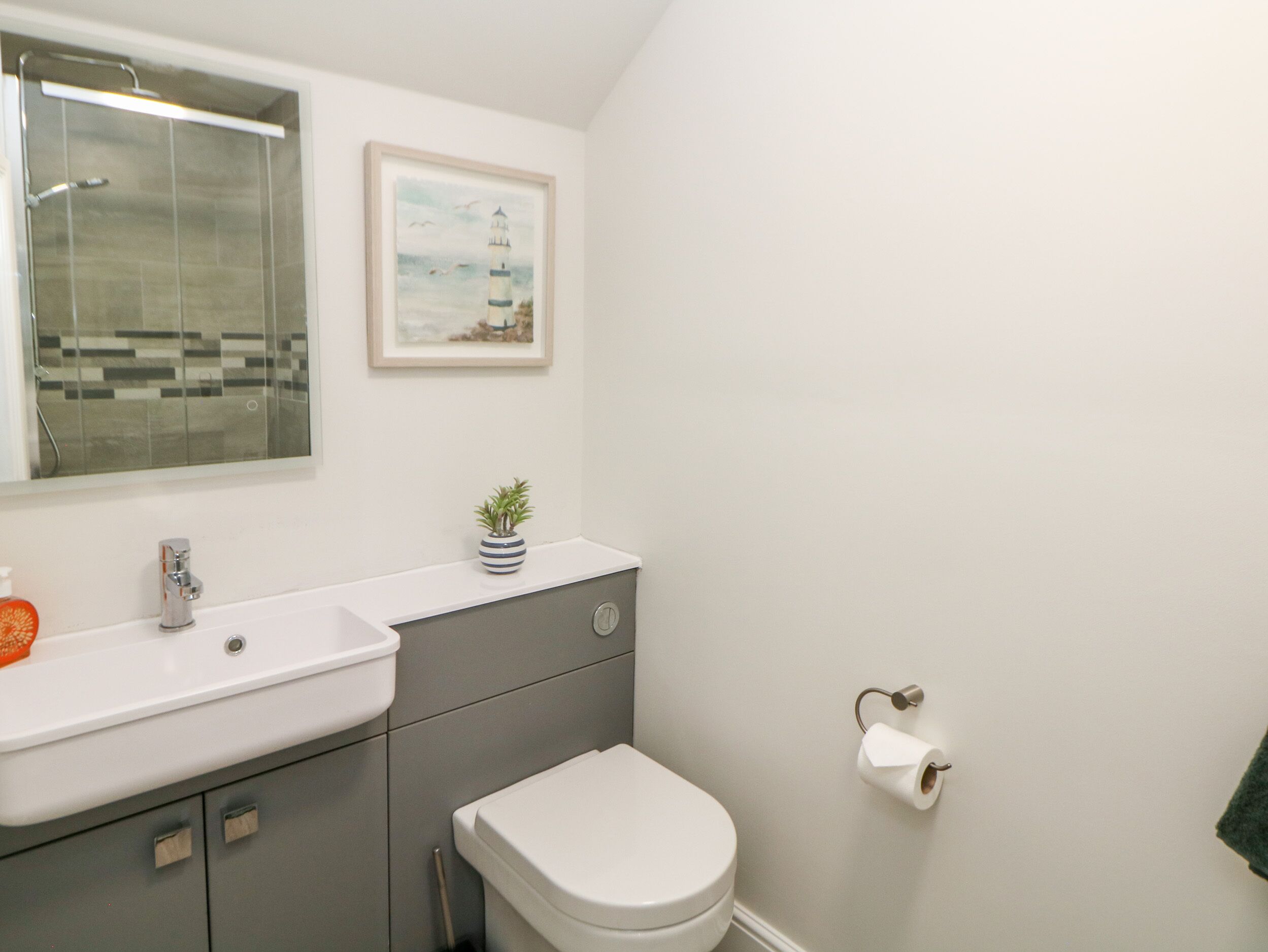Ein Badezimmer mit einem weißen Waschbecken, einer Toilette, einem kleinen Topfpflanzen, einem gerahmten Bild von einem Leuchtturm und einer Duschkabine aus Glas.