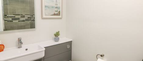 Ein Badezimmer mit einem weißen Waschbecken, einer Toilette, einem kleinen Topfpflanzen, einem gerahmten Bild von einem Leuchtturm und einer Duschkabine aus Glas.