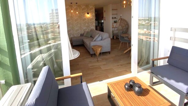 Ein Balkon mit einer Couch, einem Holztisch mit Dekoration und Blick auf eine Stadt durch große Glasfenster.
