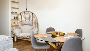 Ein Essbereich mit einem runden Glastisch, grauen Polsterstühlen, einem hängenden Rattan-Sessel und einem kleinen Beistelltisch mit einer Kerze.