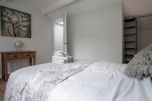 A bedroom with a bed, a bedside table with a mirror, a stack of folded towels, a painting on the wall, and a closet with shelves.