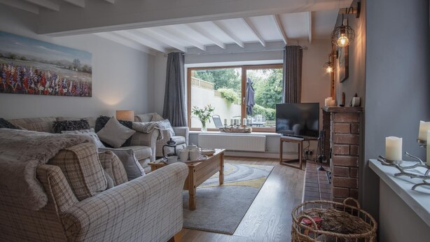 A cozy living room with a sofa, a coffee table, and a TV. There's a fireplace, a window with curtains, and a painting on the wall. - Wern Y Glais - 2 Bedroom Cottage - Glais (Swansea)