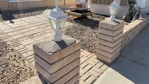 Zona residencial con un patio empedrado, dos faroles blancos de estilo exterior, una mesa pequeña y una ventana con persianas.