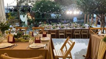 Un área de comedor al aire libre con mesas dispuestas para una comida, rodeada de sillas y decorada con plantas.