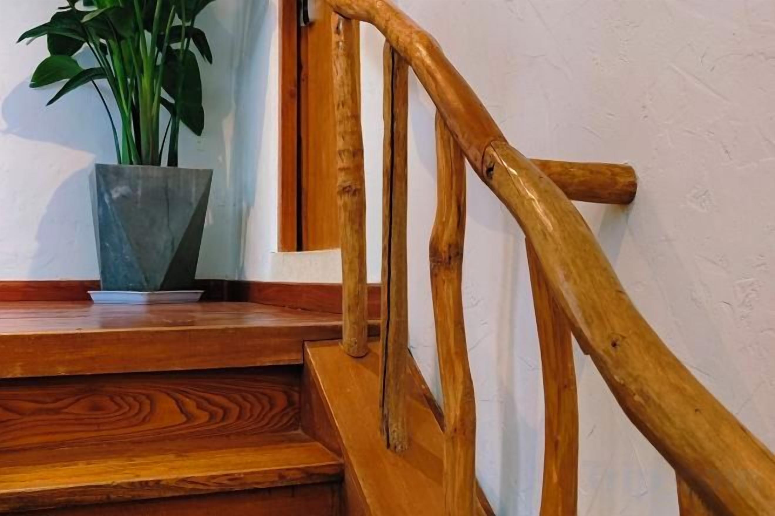 Una escalera de madera con barandilla, una planta en maceta en el rellano y una pared blanca.
