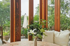 Una terraza de madera con un sofá blanco y un florero, rodeada de altos pilares de madera y follaje.