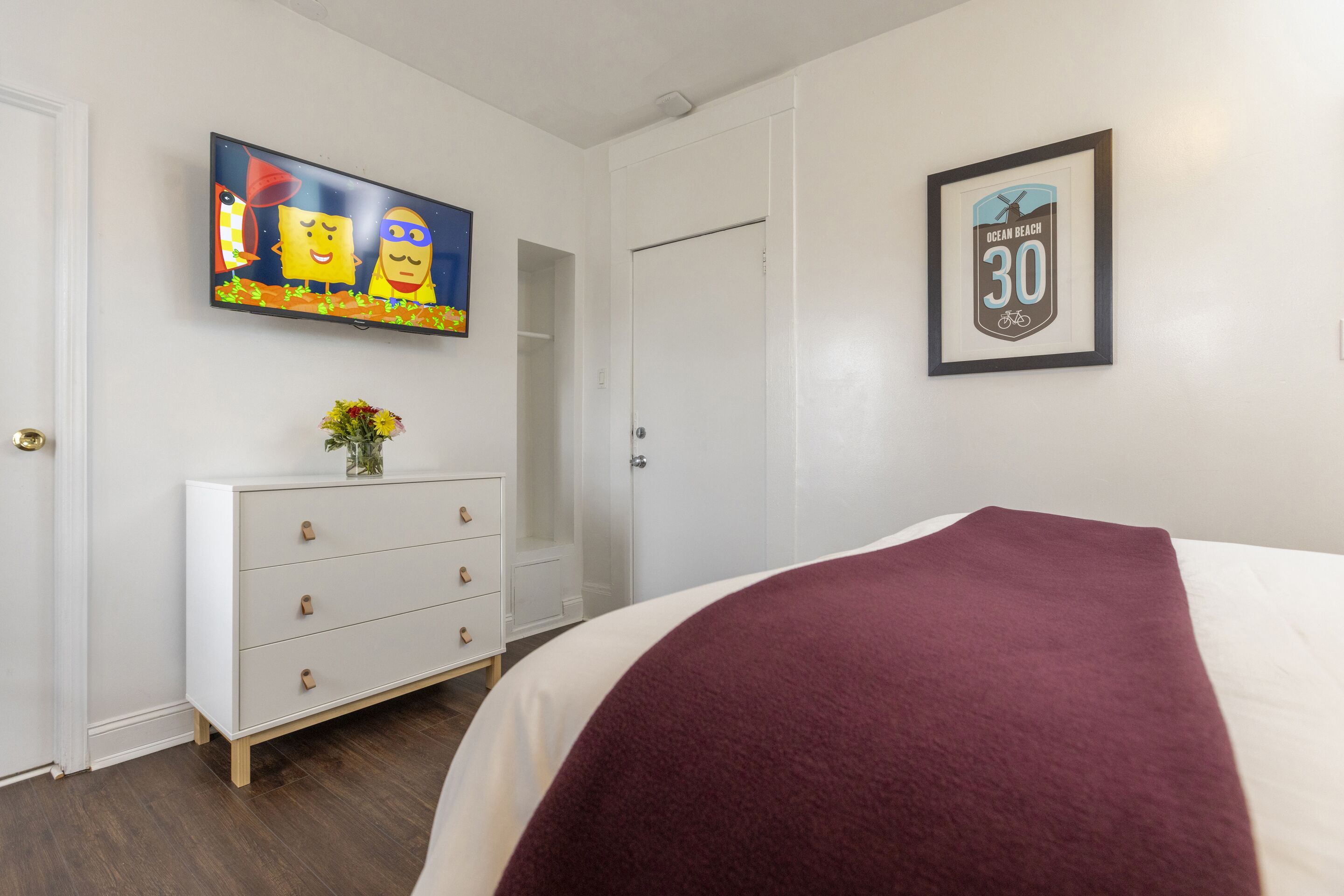 Een slaapkamer met een bed, een dressoir, een aan de muur bevestigde televisie en een lijst met een bordje.